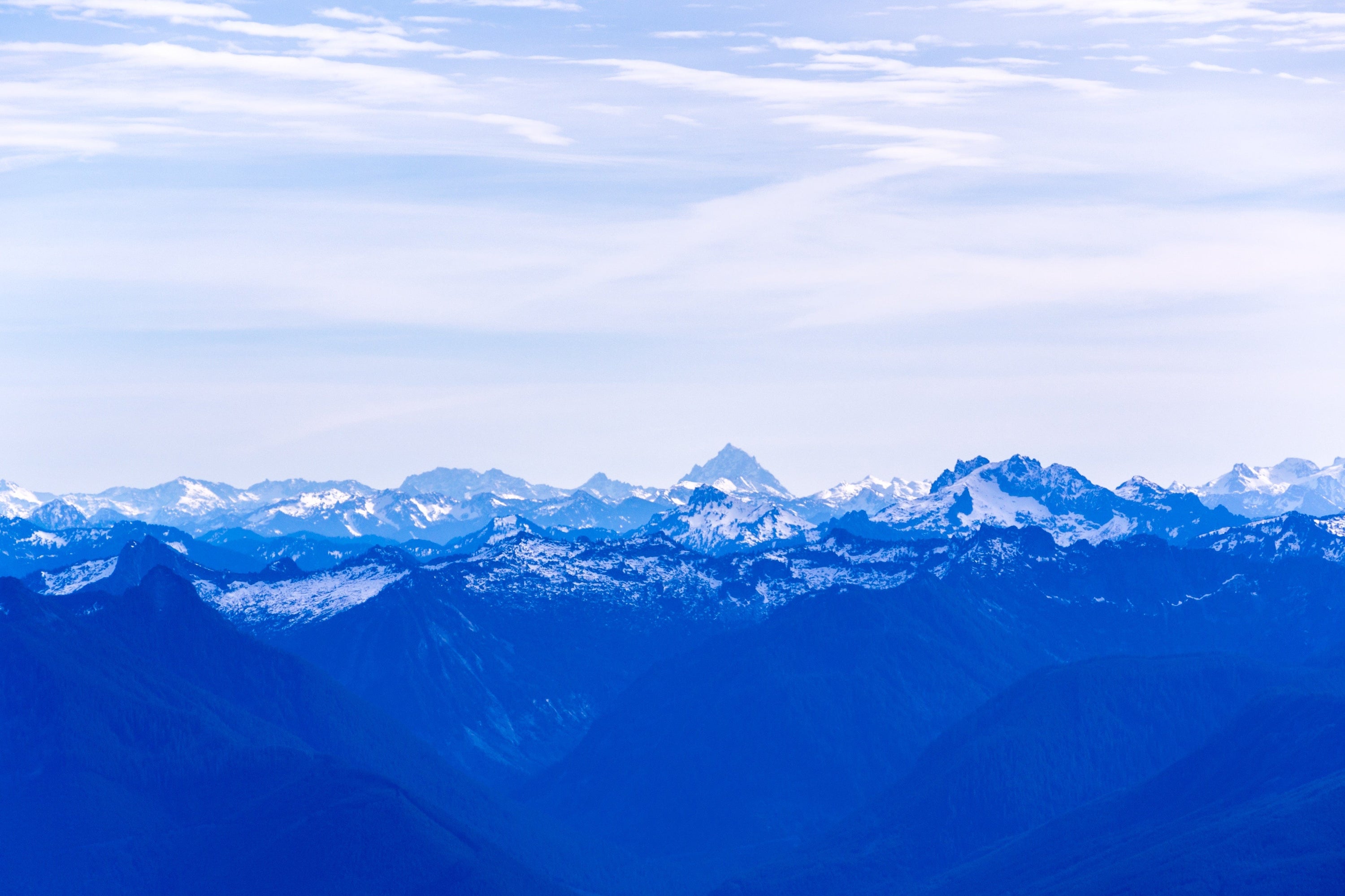 The hazy blue color of the sky is out-matched by the deeper blue of the mountains below. Stretching off into the distance, these seemingly never-ending peaks look sharp and scattered with snow.