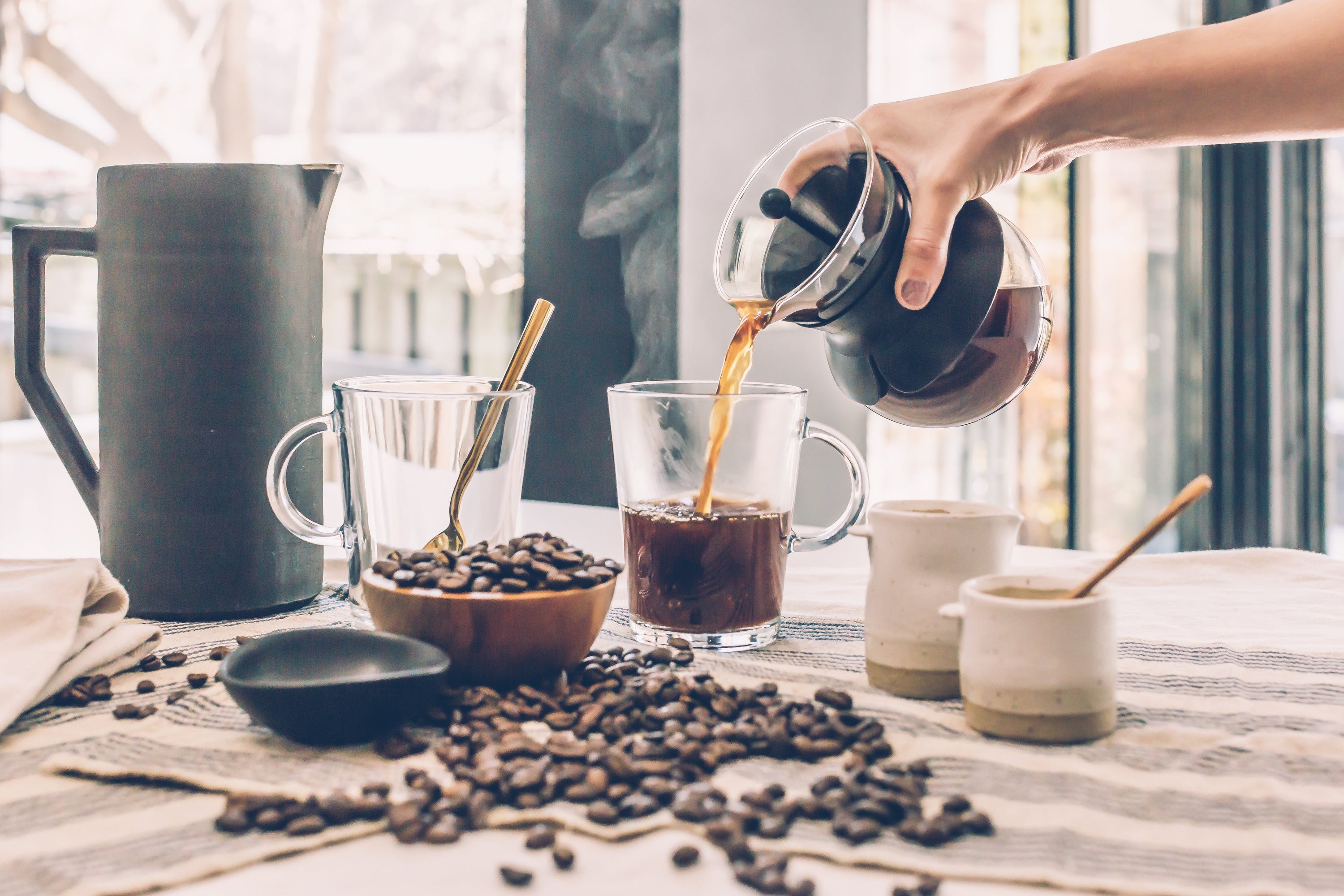 Table with French press coffee being poured into one of two glass mugs with sugar and creamer containers and spilled roasted coffee beans