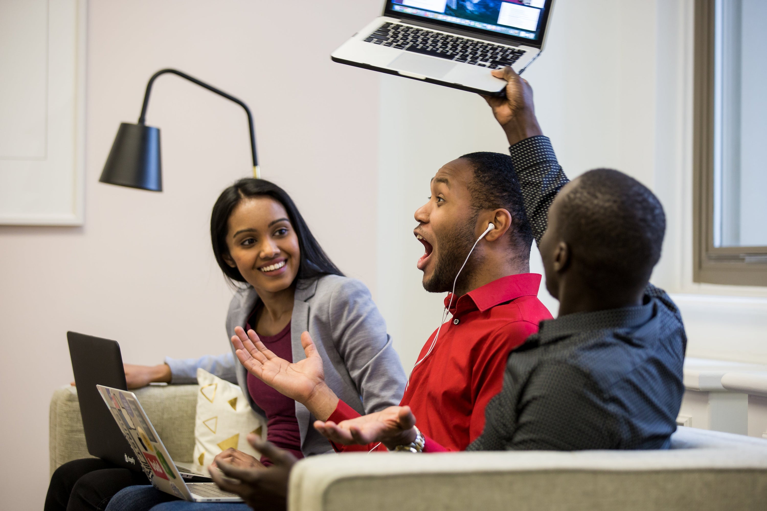 Three young professionals are sitting on a couch working on their laptop computers. They are discussing something together