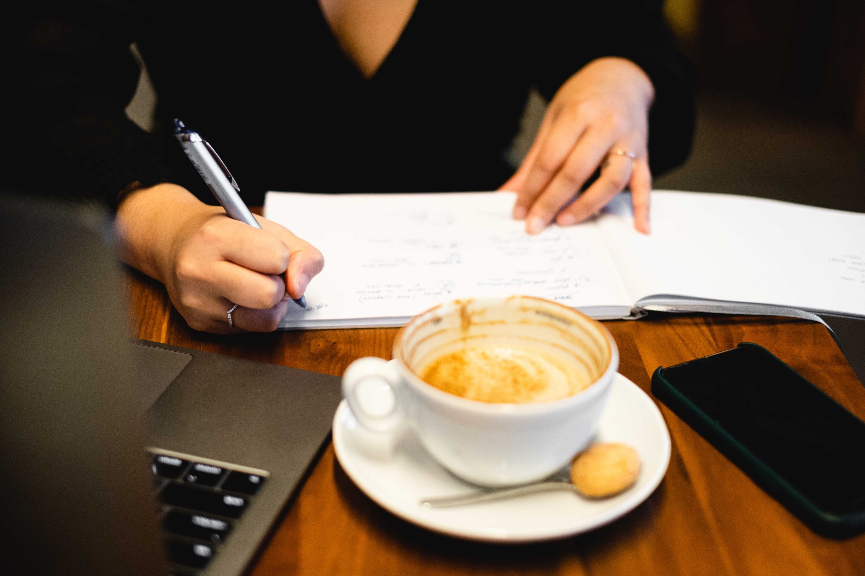 View of a laptop an empty coffee cup and a cell phone sit in front of hands writing in a open notebook