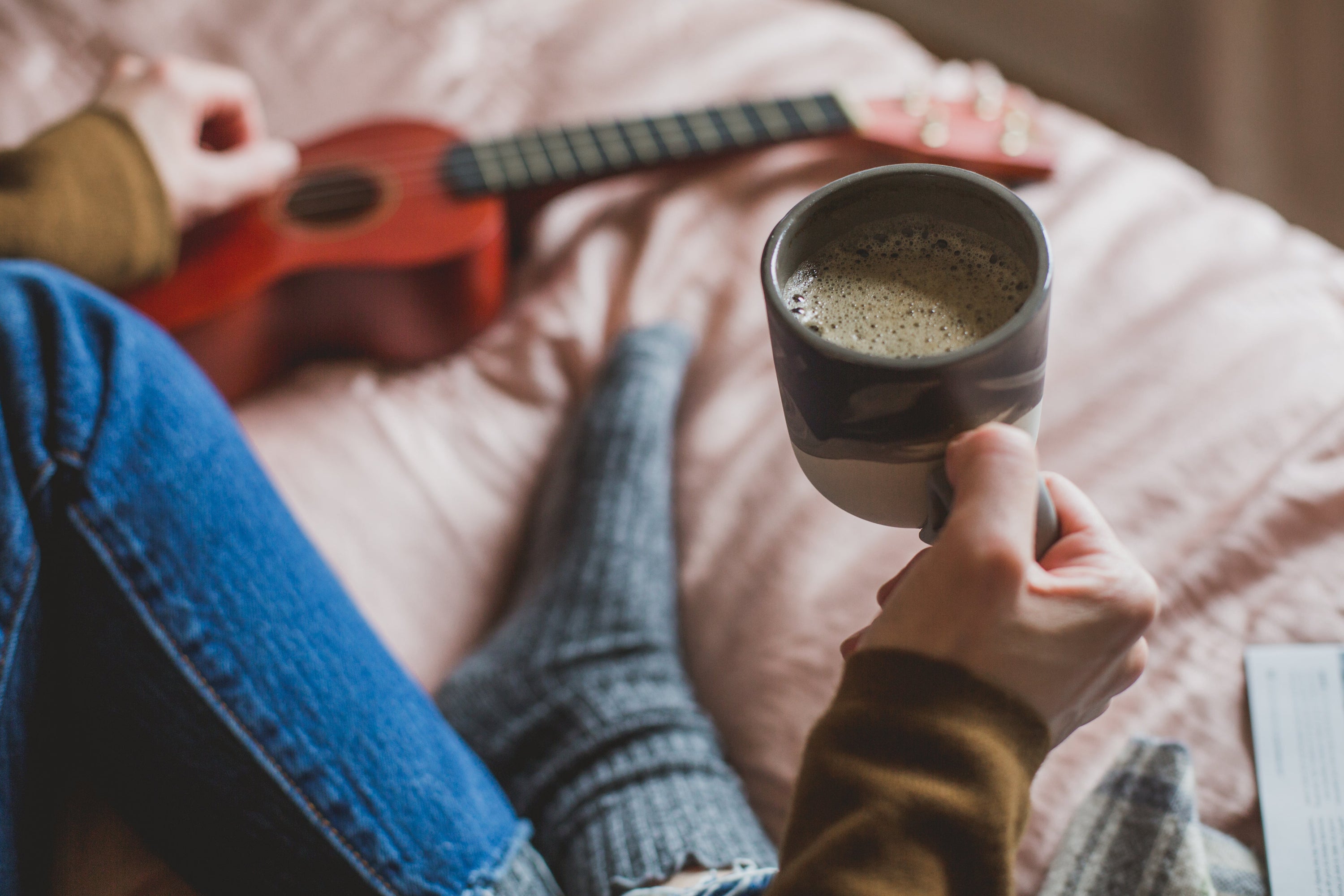 A person sits cross-legged on a bed. In the frame are one leg wearing blue jeans, one foot wearing a sock, and two arms wearing yellow sleeves. The right-hand holds a full cup of coffee, the left hand plucks at a ukelele lying flat on the blanket.