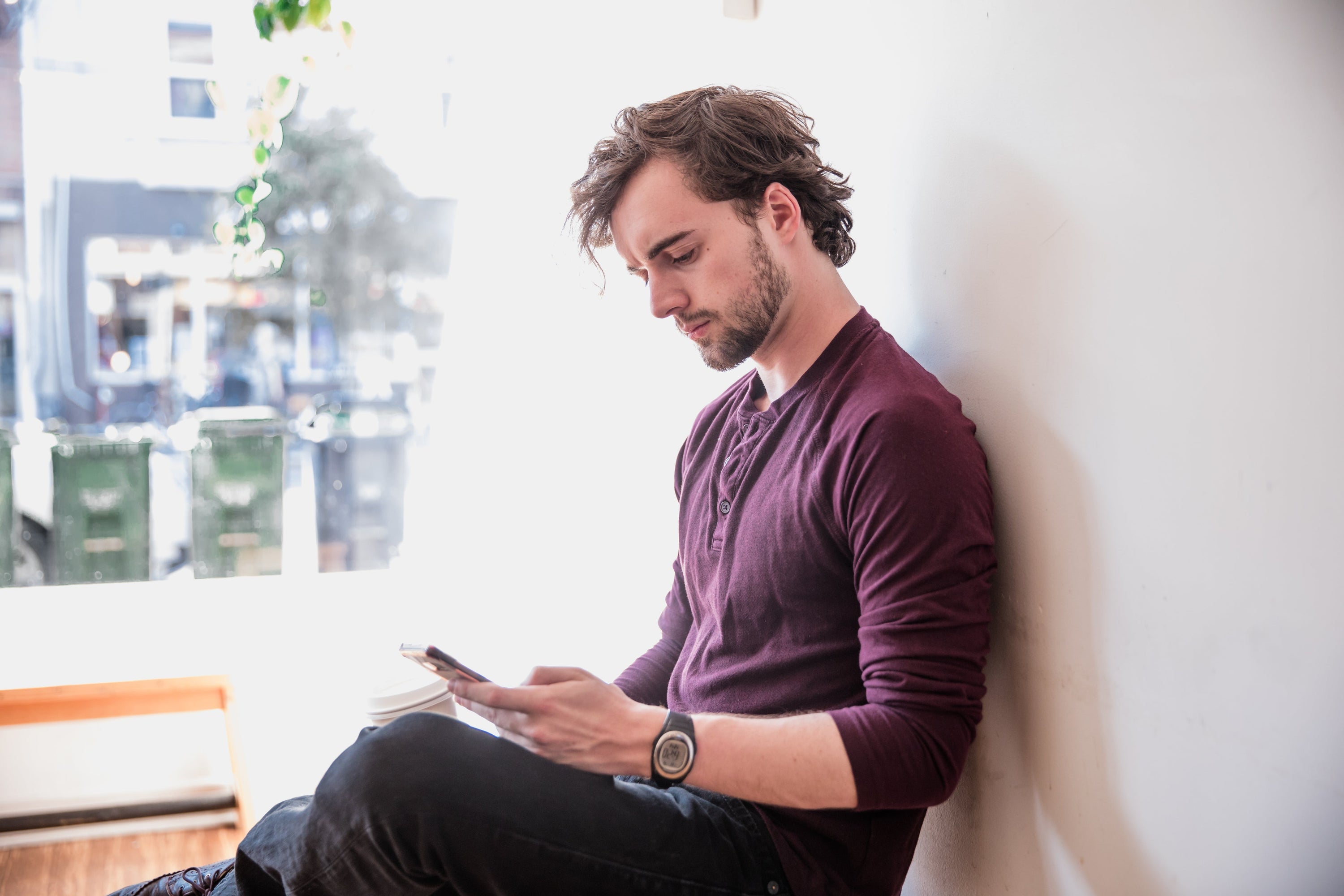 A man sits by a window looking thoughtfully into his mobile device as he goes through the days emails.