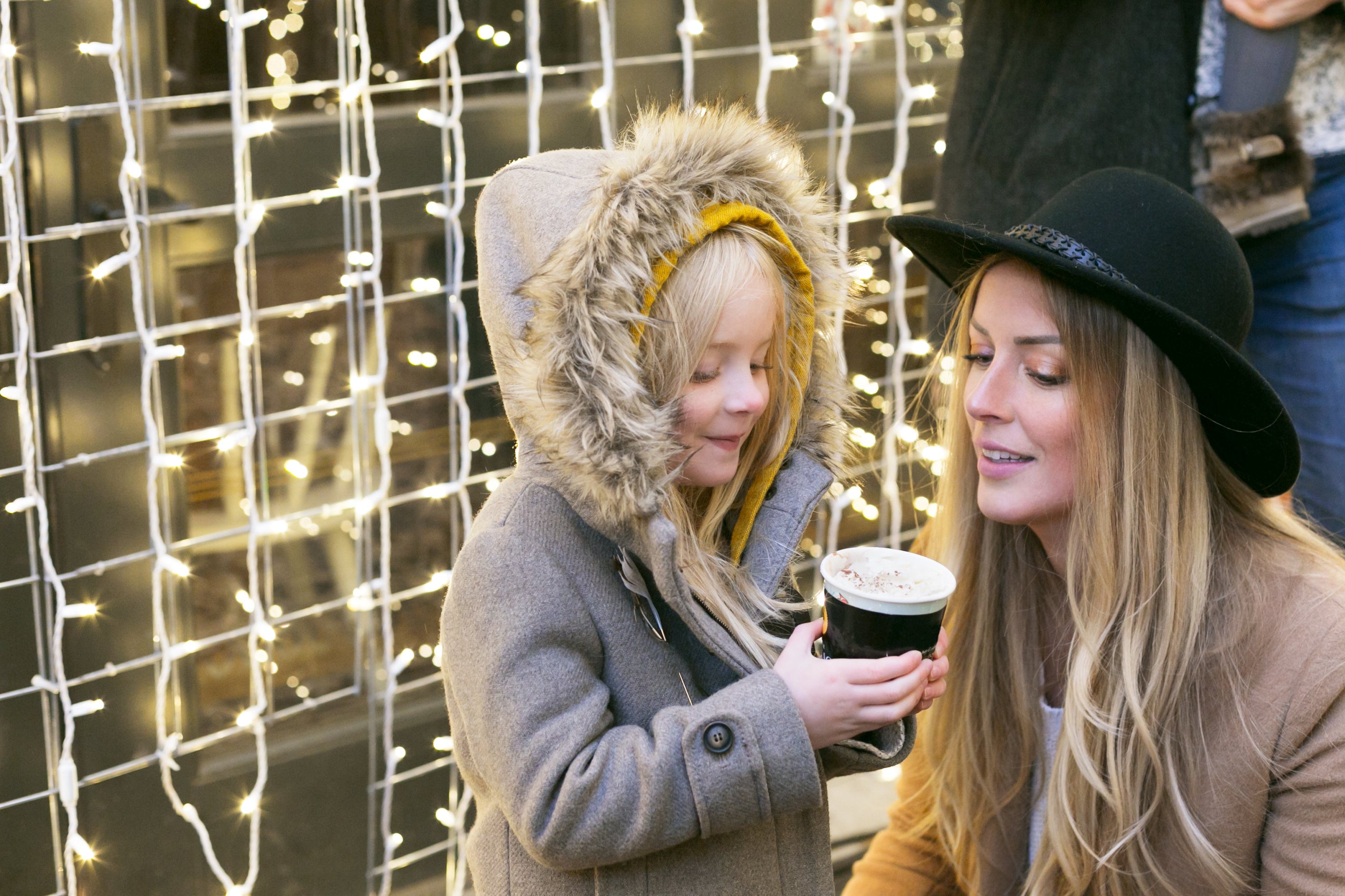 A little girl holds a cup of hot chocolate at the Christmas market with her mother in front of a wall of stringed lights.