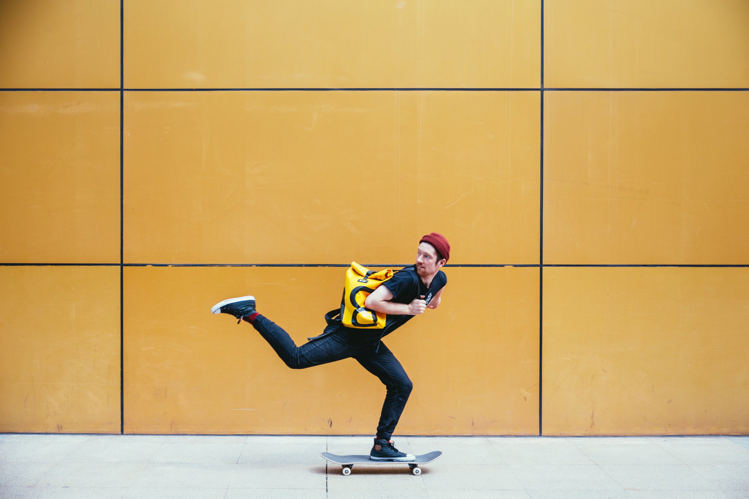 A skateboarder kicks their leg high and looks behind them as they ride past a yellow wall.