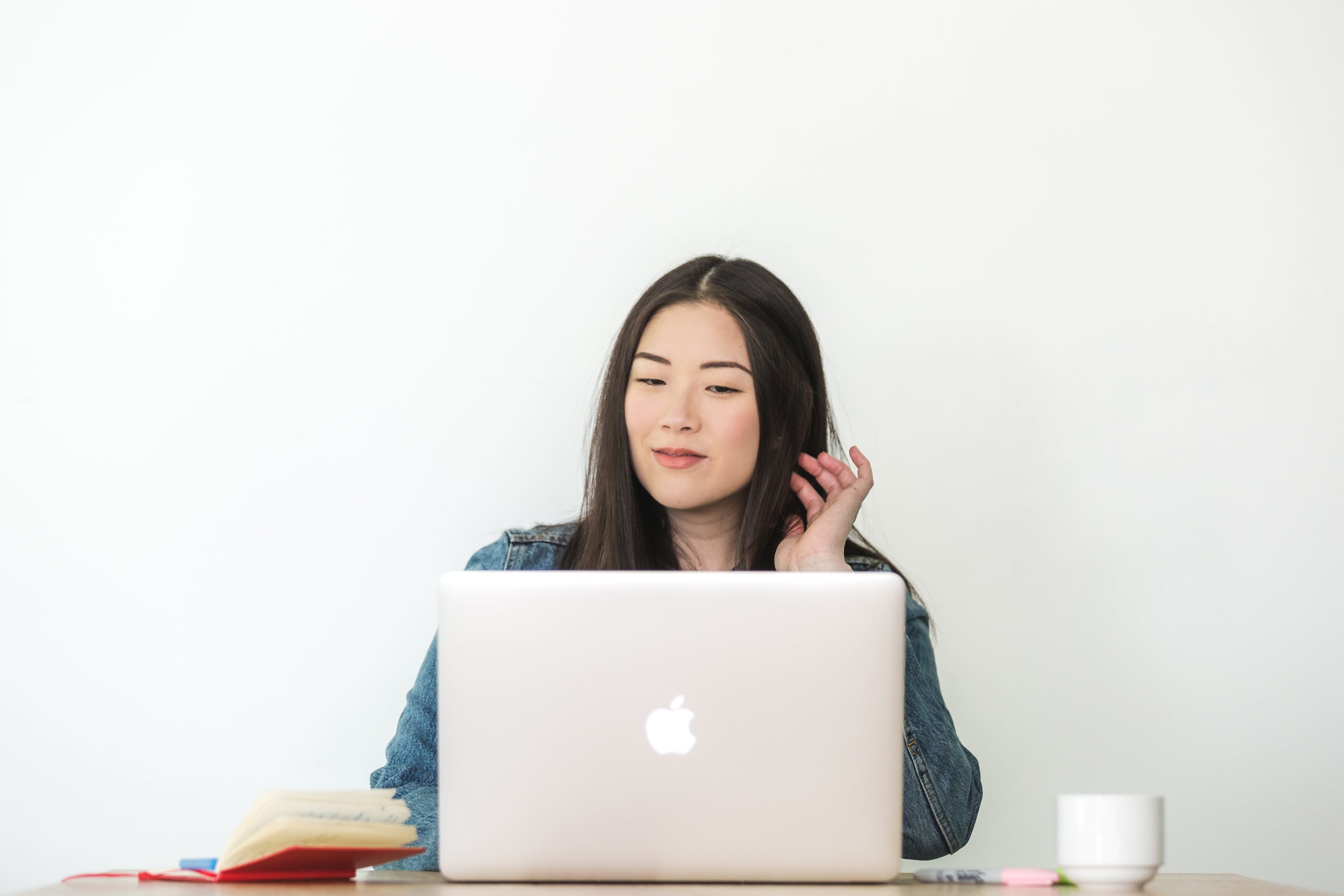 A woman sits working on a laptop with her coffee and a notebook.