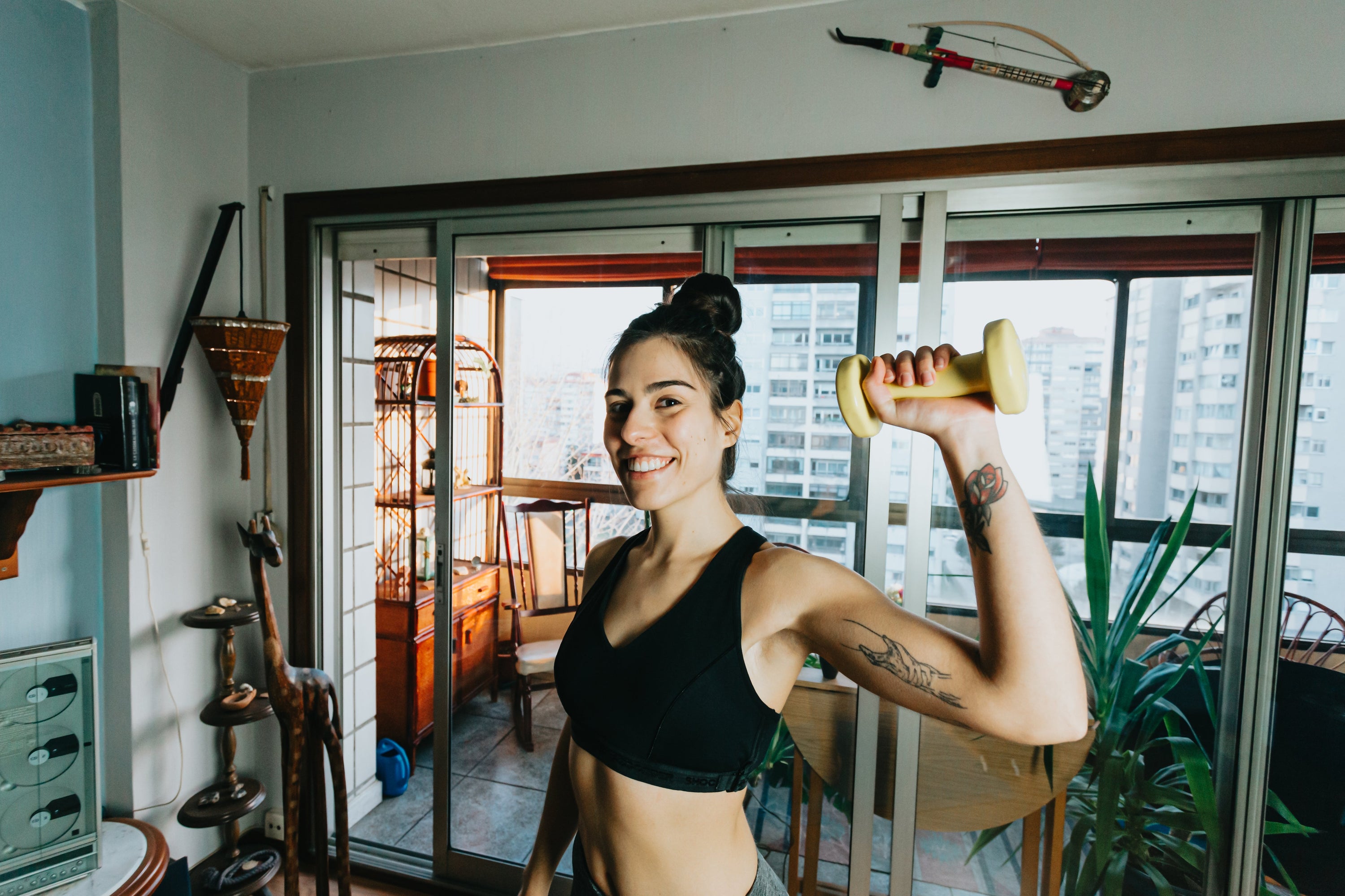 Woman smiles at the camera and holds a weight up with one hand while standing in their living room