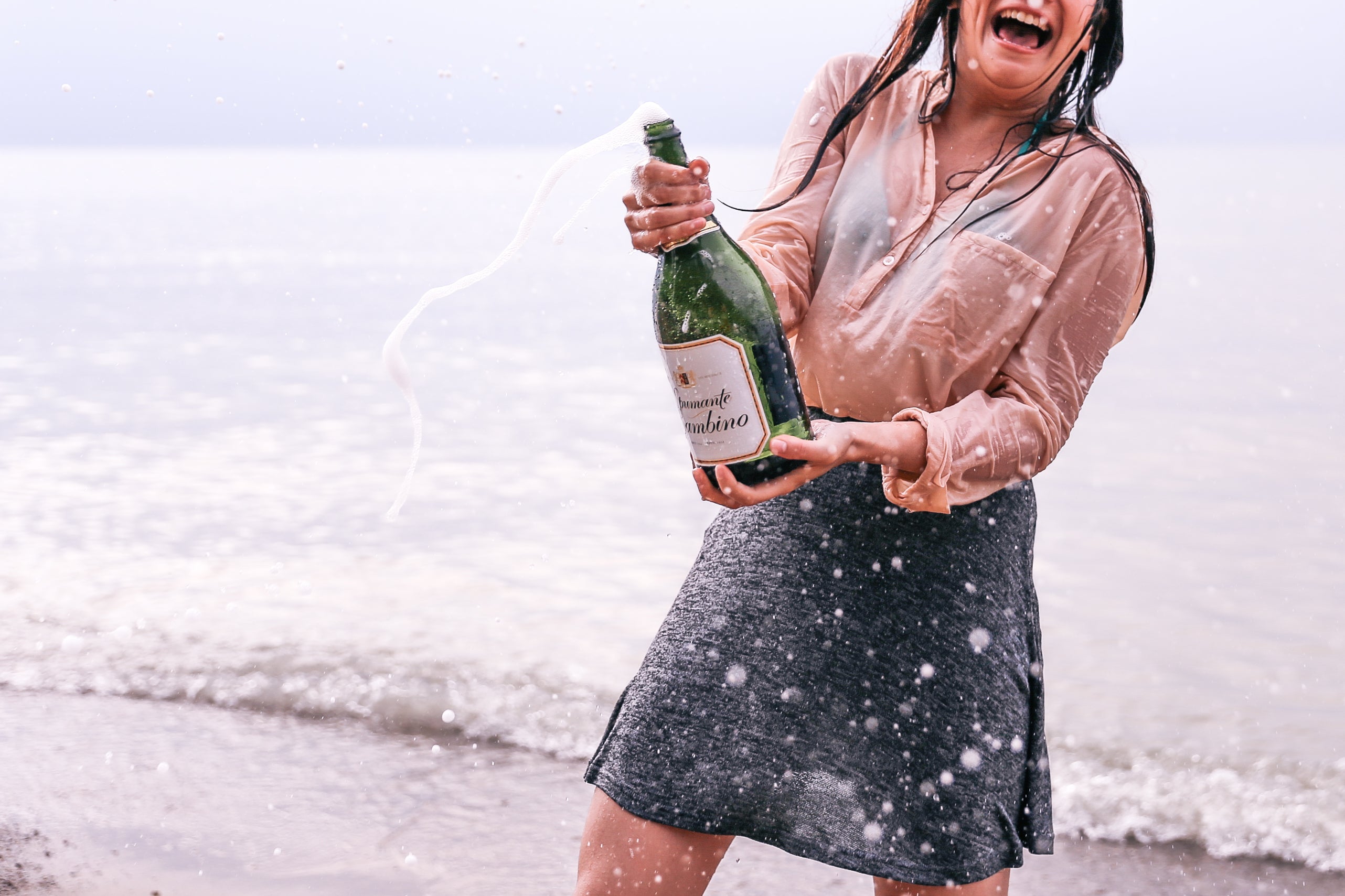 An excited woman opens a bottle of champagne on the edge of a beach.