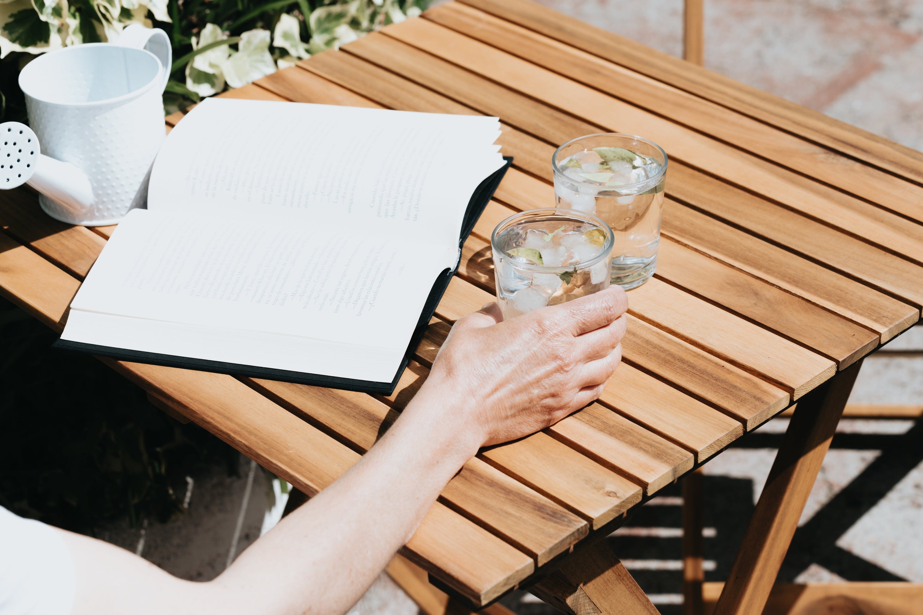 The camera looks down towards a wooden table with a open novel and two drinks. There is a hand holding one of the two drinks.
