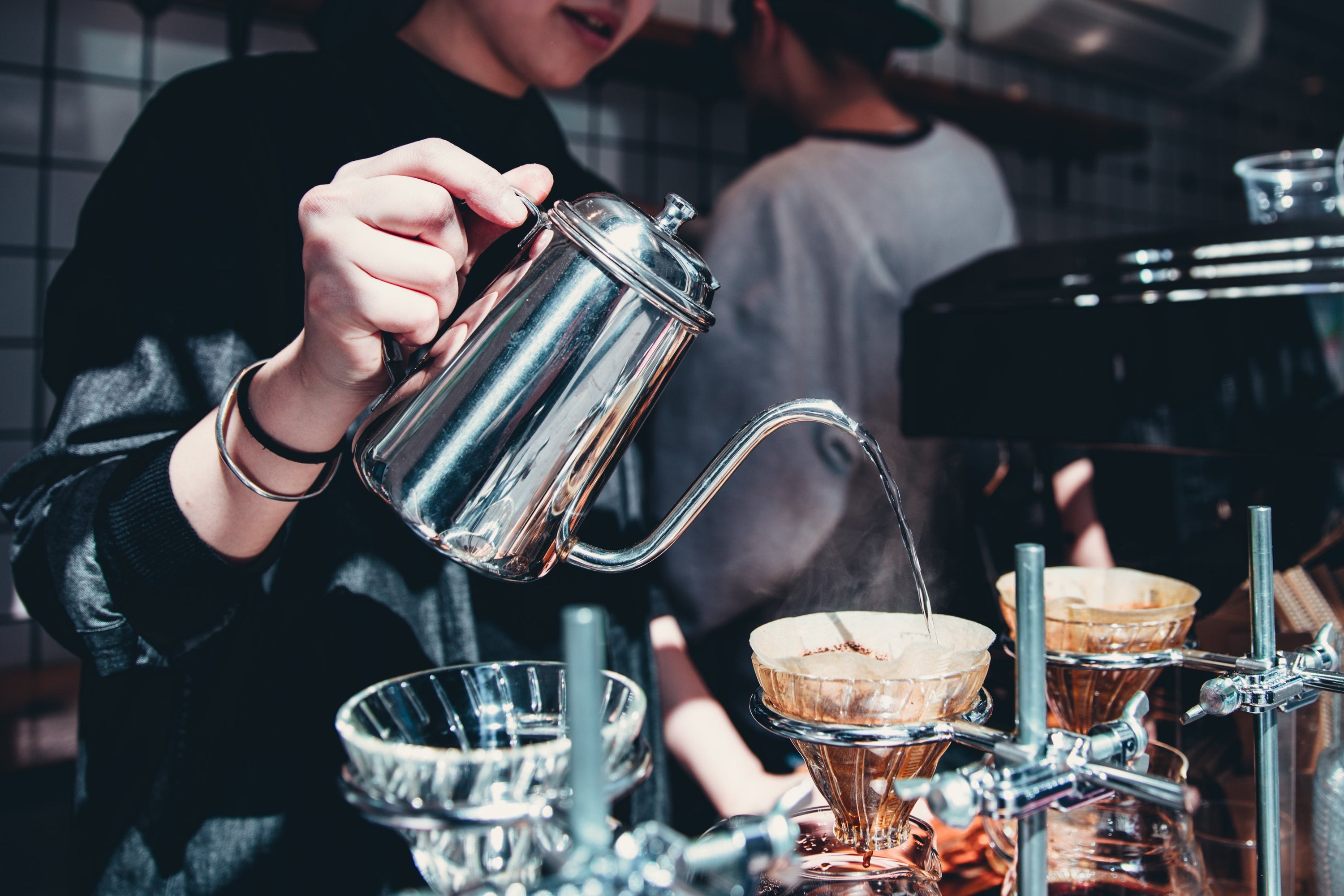 Barista pours over coffee from a steel kettle in a busy coffee shop