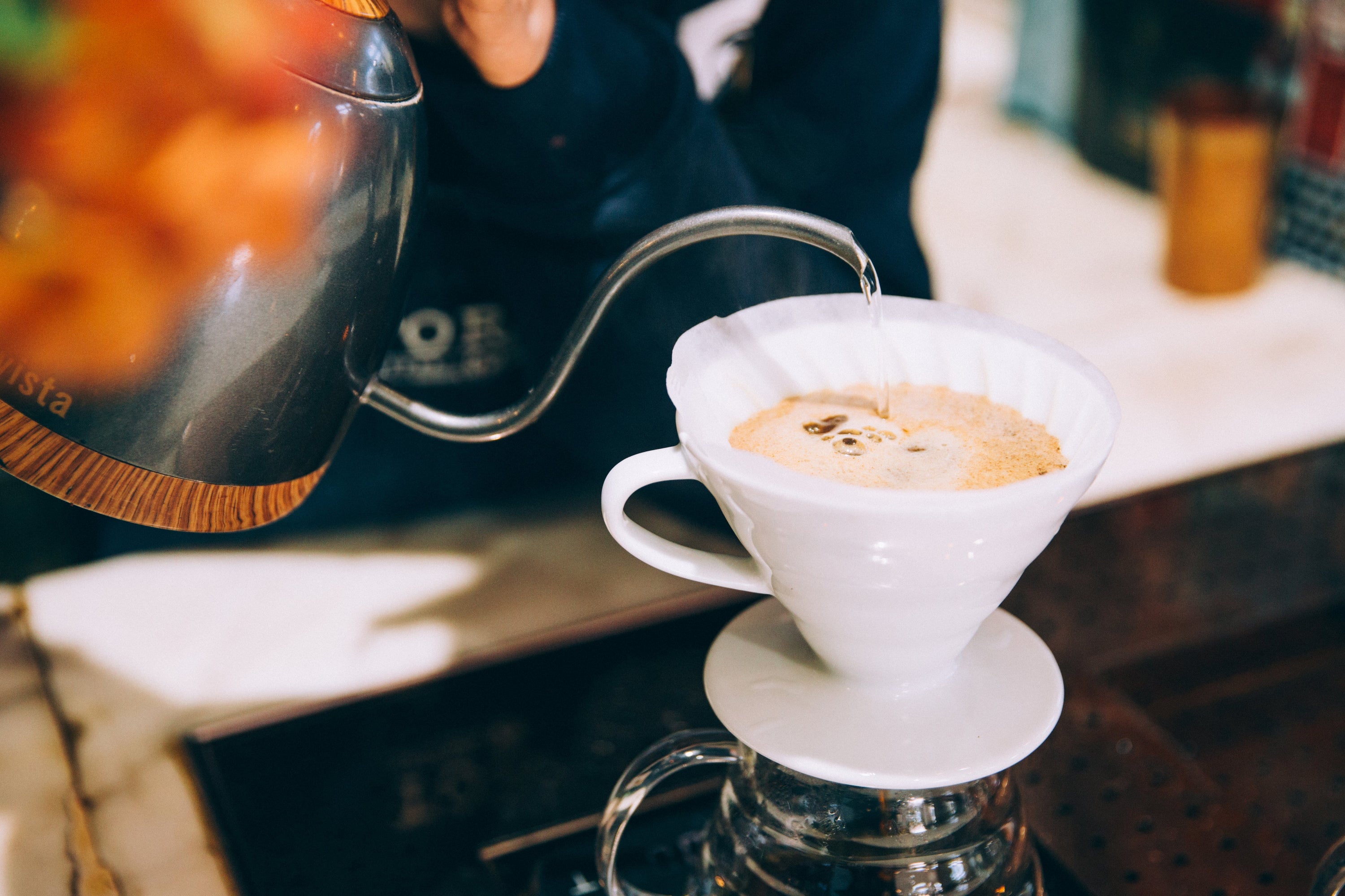 A barista carefully pours hot water from a long spout into a cup with a coffee filter.