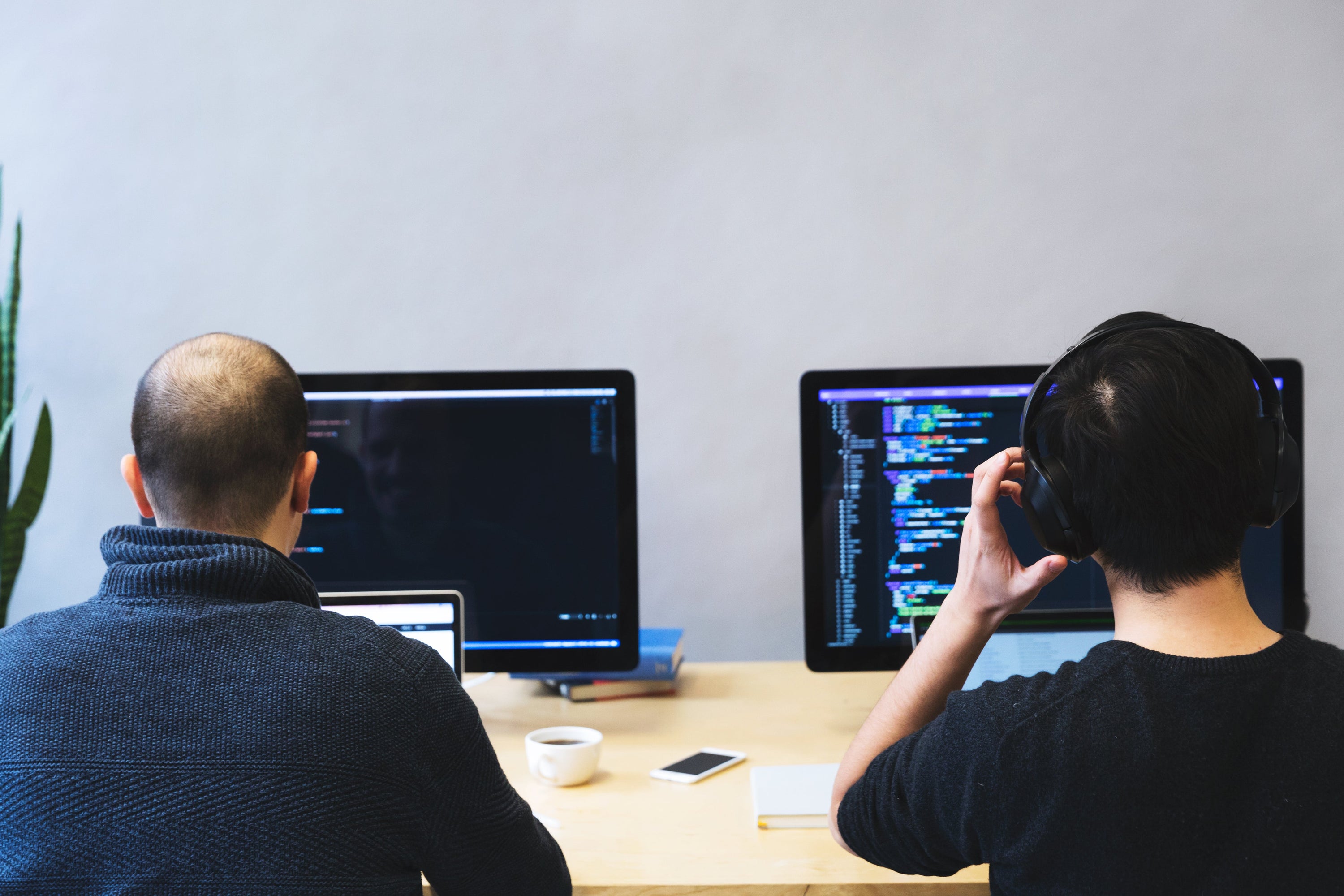 Two developers sit in front of their laptops and code in Javascript. You can tell they're in focus mode as they've got their headphones on and coffee within arm's reach