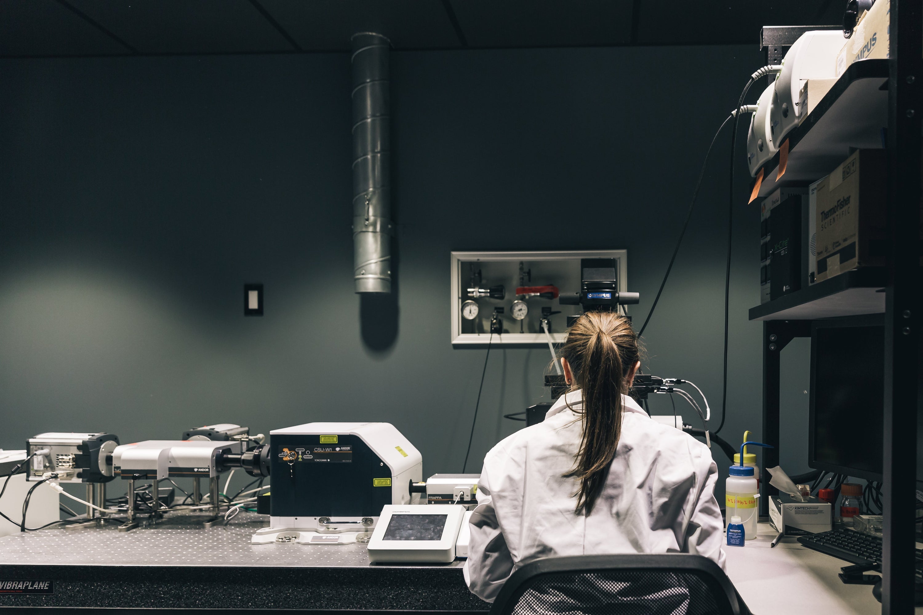 The back of a woman scientist seated at a desk in her lab, with machinery on the shelves to the right and all along the desk in front of her. Dials and knobs and wires, oh my!