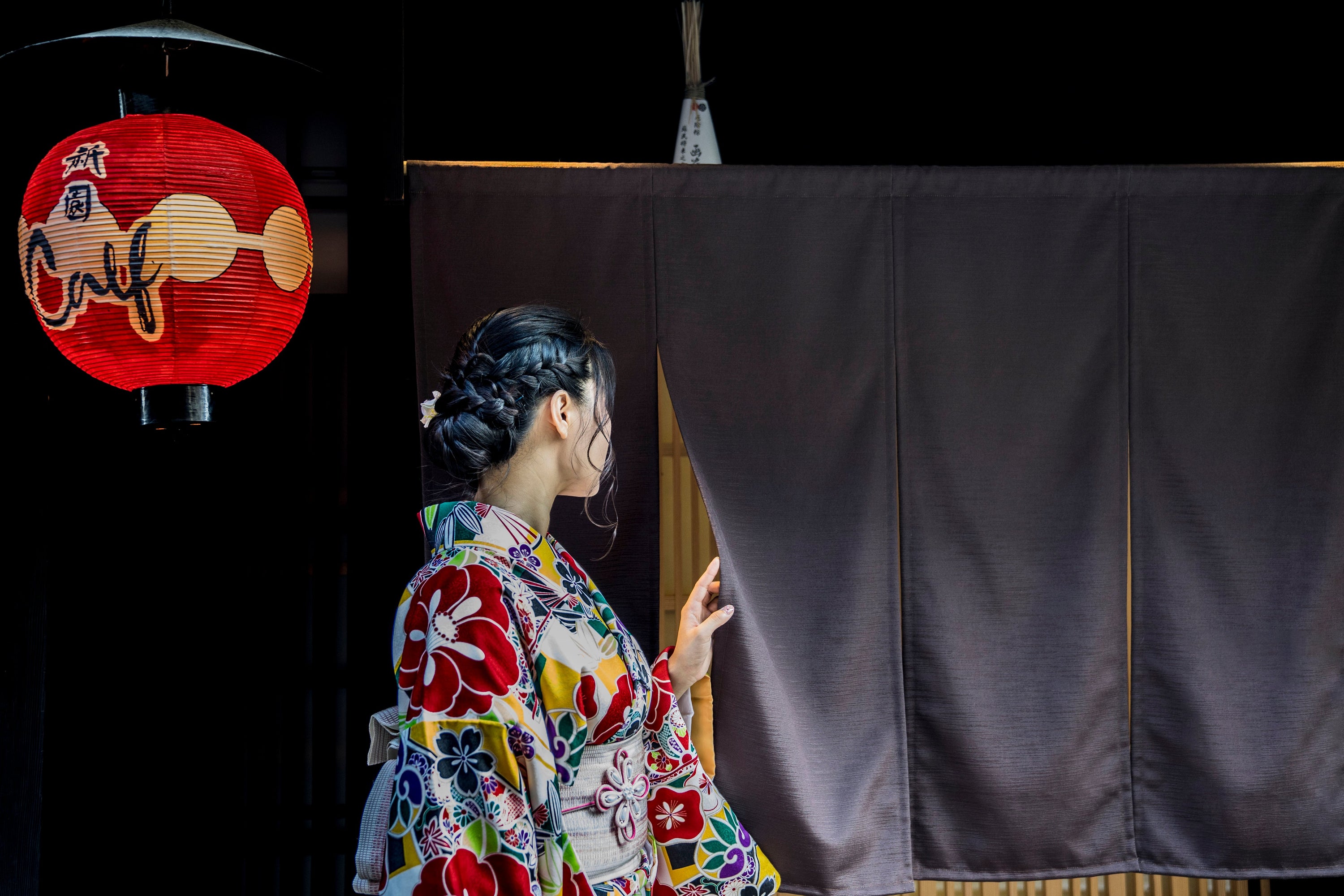 A person wearing a traditional kimono peaks behind a black curtain. There is a red paper lantern behind them.