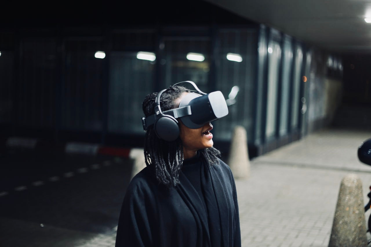 Person stands in the middle of the frame and wears a VR headset covering their eyes. Behind them is pavement and building windows.