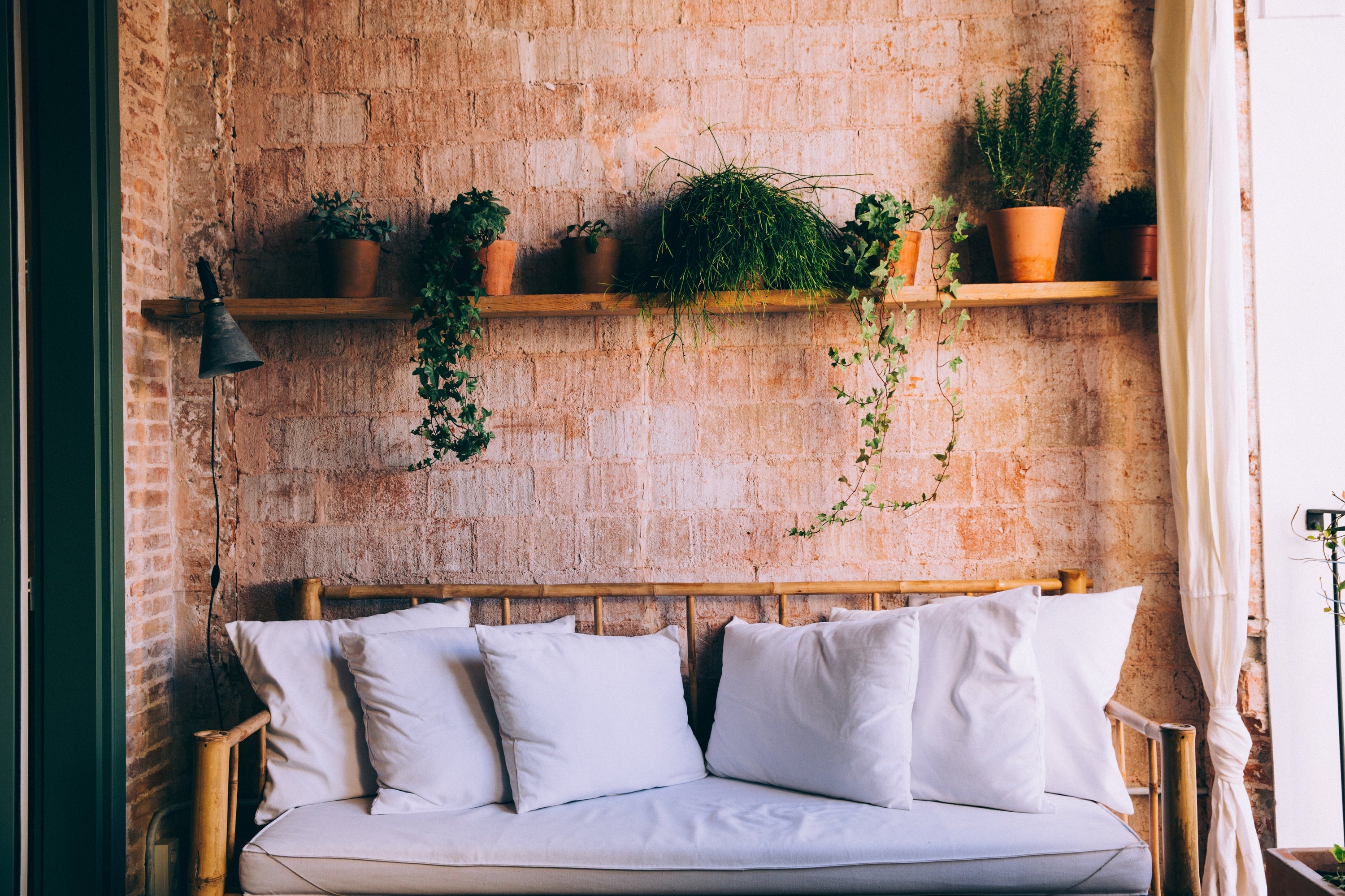 This is the perfect little nook to sit and read a book, or even have a quiet cup of coffee. A rustic wicker couch has fluffy white cushions on it, and above a wooden shelf is home to a line of happy pot plants. Sunlight streams in the window to the right.