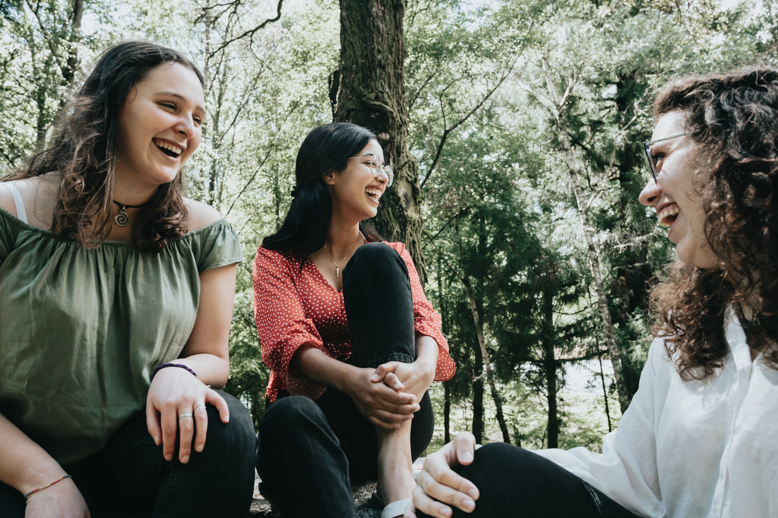 Three people sit together under a large tree in a forest. All three people are laughing and smiling wide.