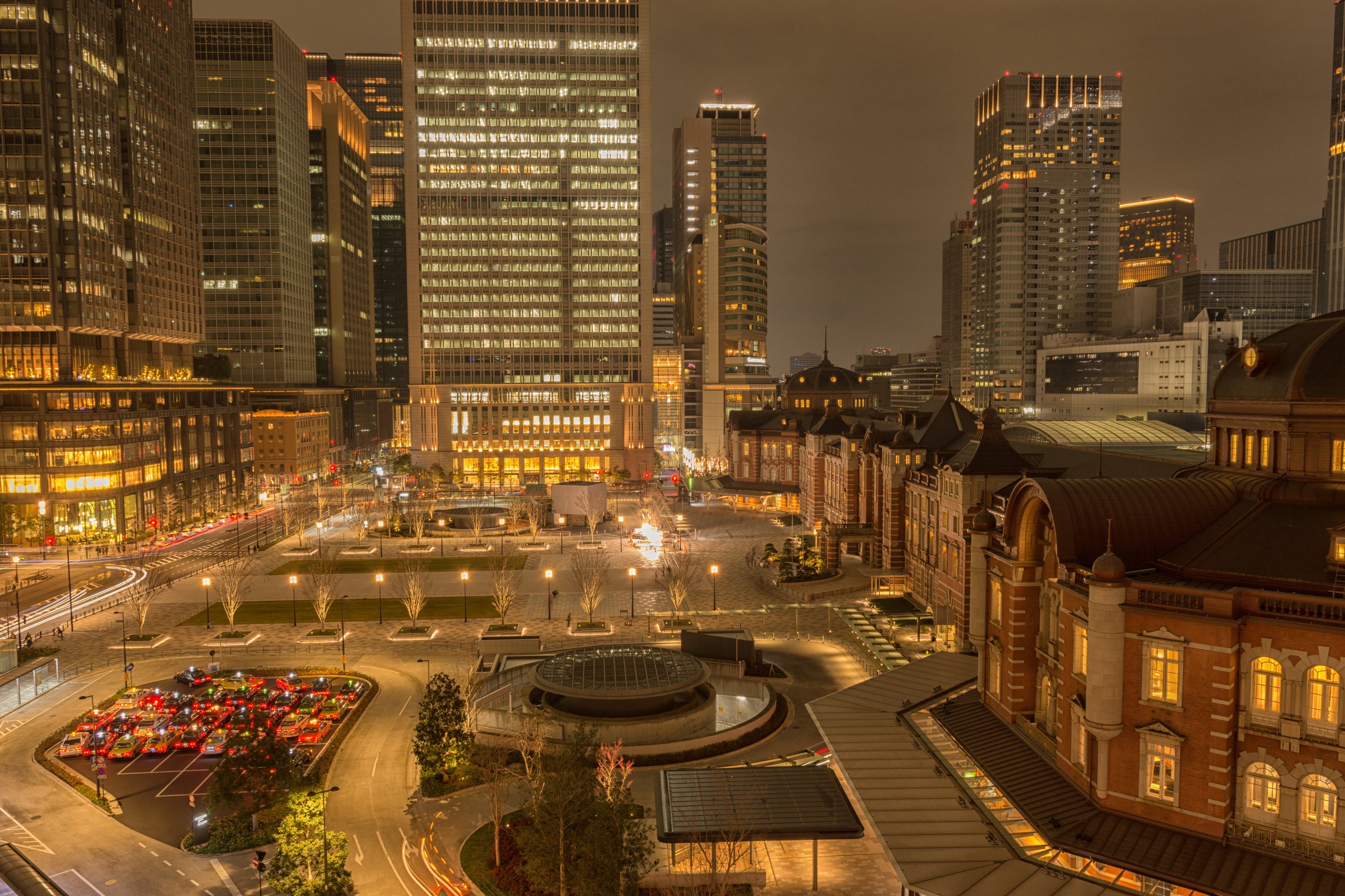 A group of lit taxi cabs waits in a quiet urban square at night, lit with the orange glow of sodium-vapor streetlights.