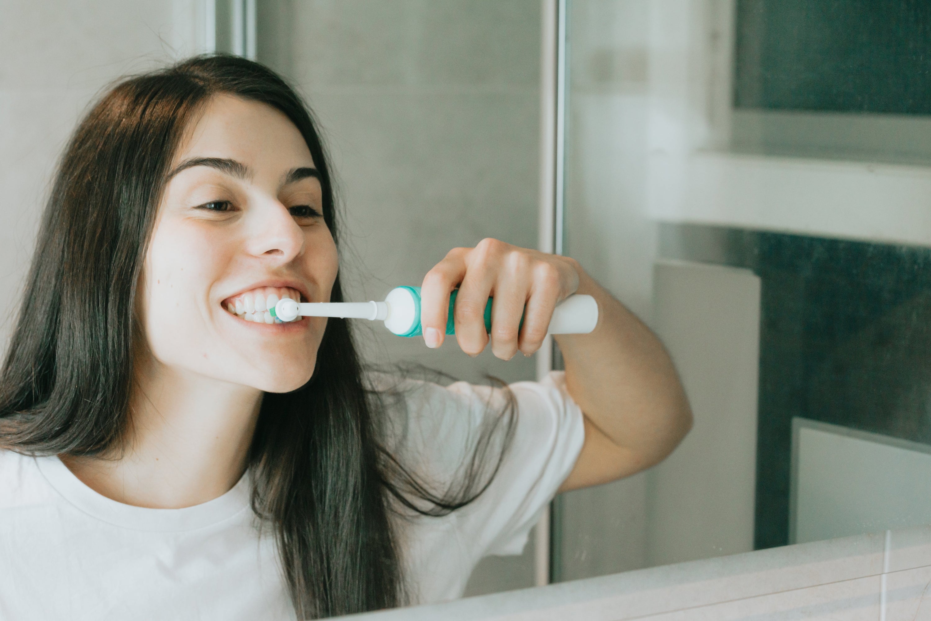 A woman looks in the mirror as she brushes her teeth with an electric toothbrush.