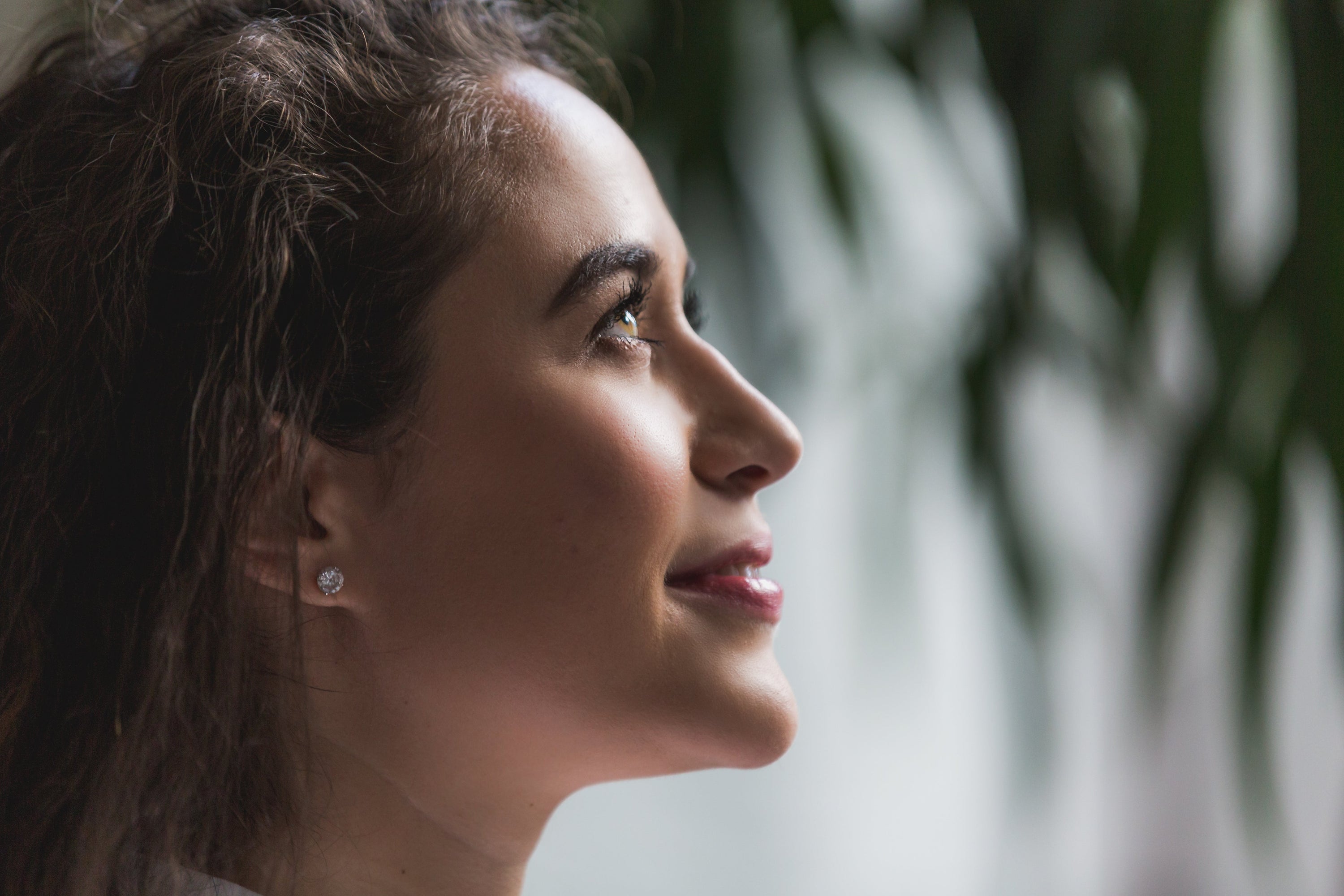 Profile of a woman's head. She has a jeweled earing and is looking up and smiling at something. Out-of-focus plans are in the background.