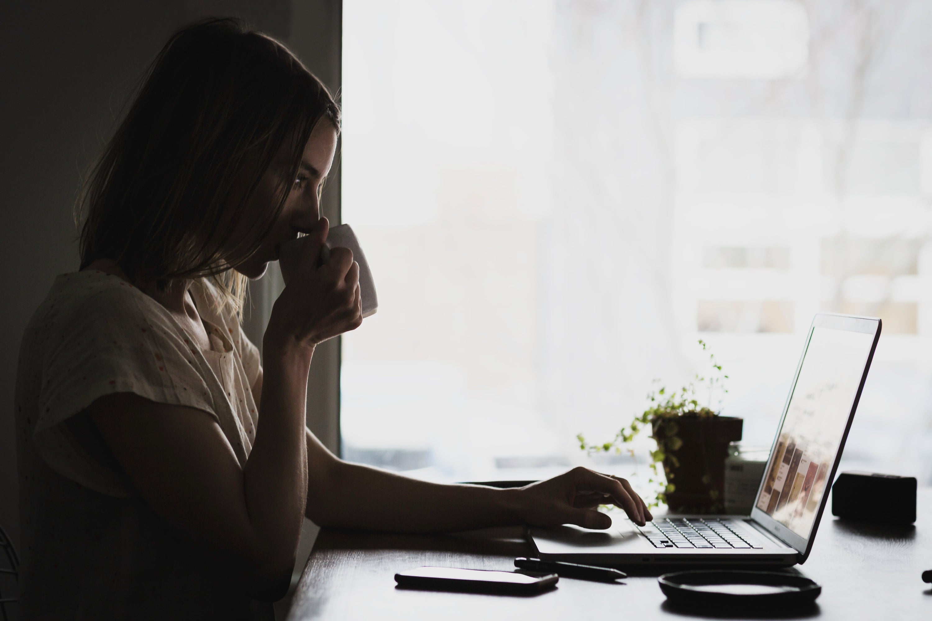 Woman sipping from coffee cup while working on laptop at desk.